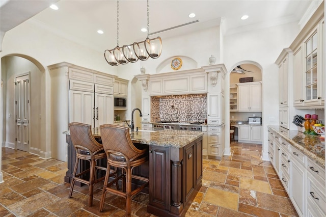 kitchen with a center island with sink, stone tile flooring, glass insert cabinets, a sink, and a kitchen breakfast bar