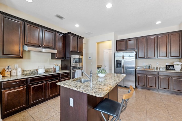 kitchen with sink, dark brown cabinets, light tile patterned floors, appliances with stainless steel finishes, and an island with sink