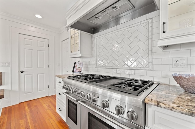 kitchen with wall chimney range hood, light stone countertops, ornamental molding, white cabinets, and range with two ovens