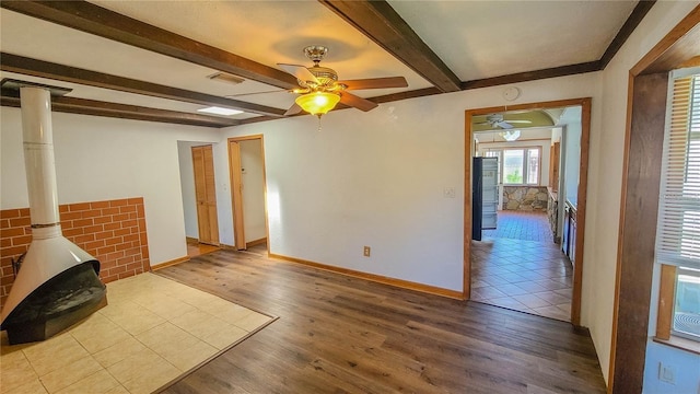 unfurnished living room with ceiling fan, a wood stove, beam ceiling, and light hardwood / wood-style flooring