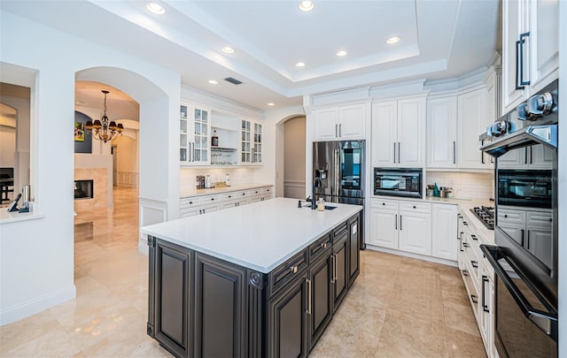 kitchen with black microwave, white cabinets, a center island with sink, decorative backsplash, and a raised ceiling