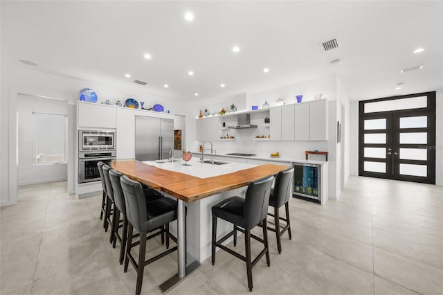 kitchen featuring beverage cooler, built in appliances, a kitchen island with sink, and white cabinets