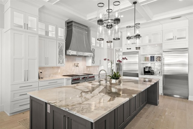 kitchen featuring wall chimney range hood, sink, white cabinetry, high quality appliances, and a large island with sink