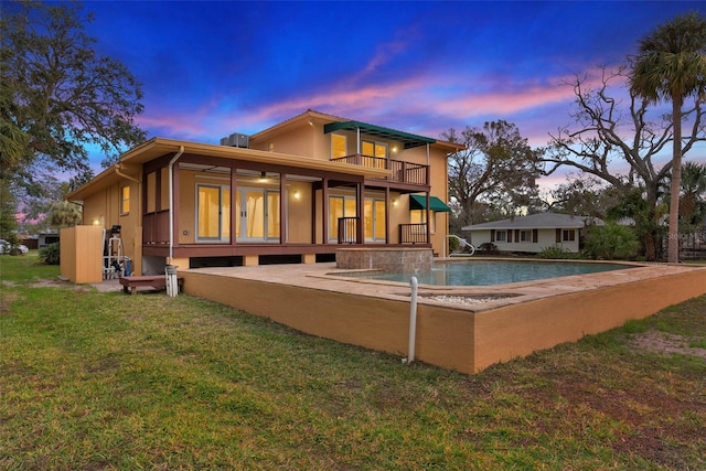 back of house at dusk with a balcony, a patio area, an outdoor pool, and a lawn