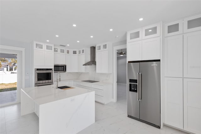 kitchen featuring wall chimney range hood, white cabinetry, appliances with stainless steel finishes, and a center island with sink