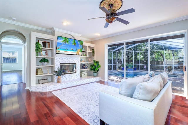 living room with a tile fireplace, built in features, dark hardwood / wood-style flooring, ceiling fan, and crown molding