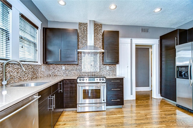 kitchen with wall chimney exhaust hood, dark brown cabinetry, sink, appliances with stainless steel finishes, and light hardwood / wood-style floors