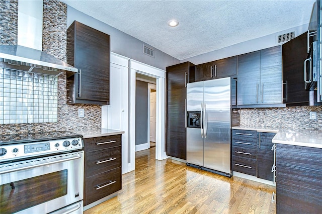 kitchen with appliances with stainless steel finishes, dark brown cabinets, decorative backsplash, wall chimney exhaust hood, and light wood-type flooring