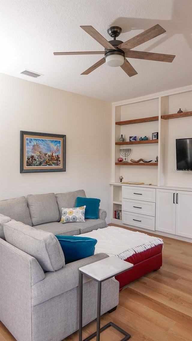 living room with ceiling fan, built in shelves, light hardwood / wood-style floors, and a textured ceiling