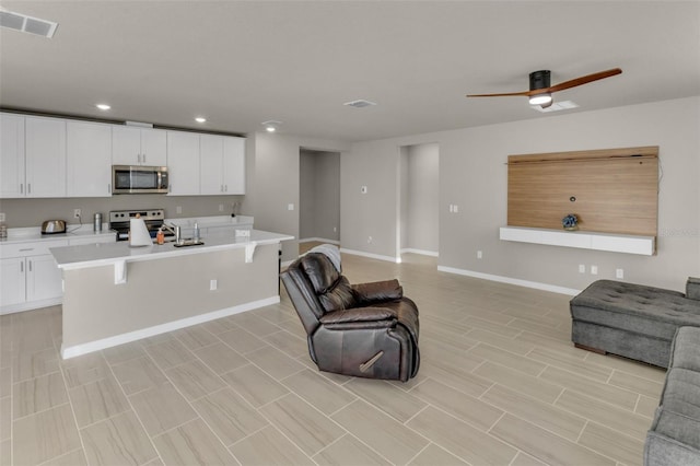 kitchen with ceiling fan, appliances with stainless steel finishes, white cabinetry, an island with sink, and a kitchen bar