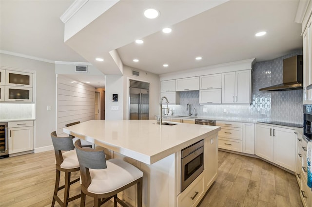 kitchen featuring white cabinetry, sink, a kitchen island with sink, built in appliances, and wall chimney exhaust hood