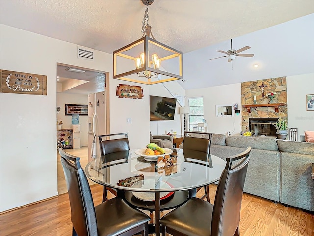 dining space featuring vaulted ceiling, a stone fireplace, ceiling fan with notable chandelier, light wood-type flooring, and a textured ceiling