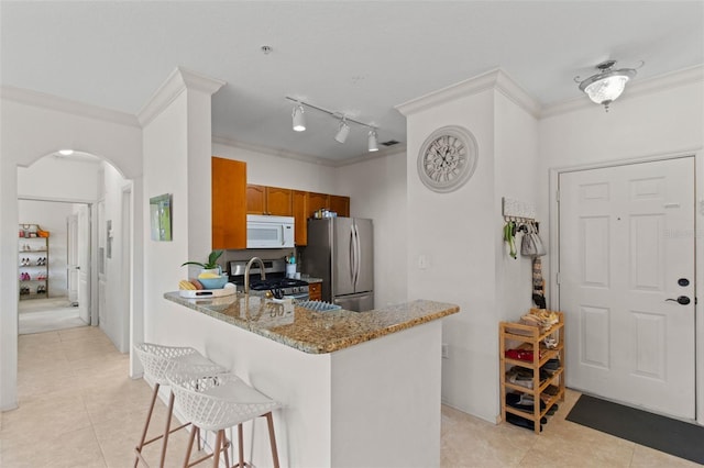 kitchen with arched walkways, brown cabinets, stainless steel appliances, ornamental molding, and a kitchen bar