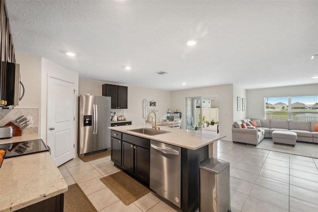 kitchen with appliances with stainless steel finishes, sink, a kitchen island with sink, light tile patterned floors, and light stone counters