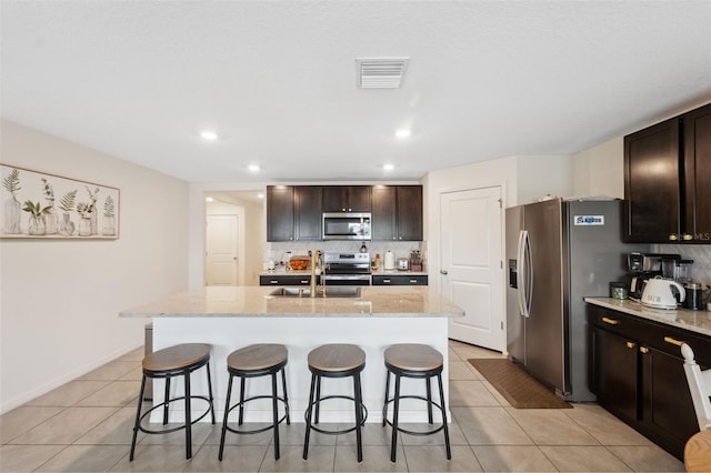 kitchen featuring an island with sink, appliances with stainless steel finishes, dark brown cabinets, and decorative backsplash