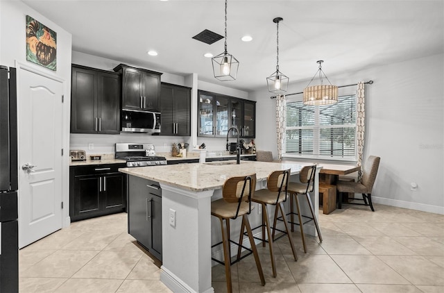 kitchen with light tile patterned flooring, sink, hanging light fixtures, appliances with stainless steel finishes, and a kitchen island with sink