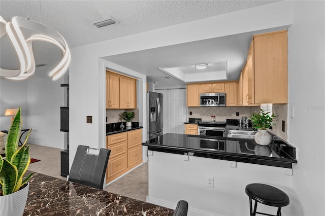 kitchen with light brown cabinetry, a raised ceiling, a kitchen bar, kitchen peninsula, and stainless steel appliances