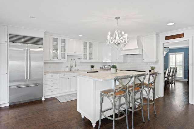 kitchen with built in fridge, premium range hood, white cabinetry, hanging light fixtures, and a center island