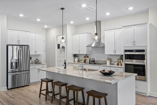 kitchen featuring white cabinetry, decorative light fixtures, a center island with sink, stainless steel appliances, and light stone countertops