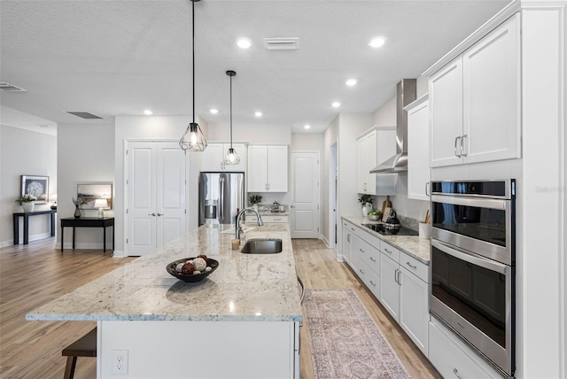 kitchen featuring wall chimney exhaust hood, sink, a center island with sink, appliances with stainless steel finishes, and pendant lighting