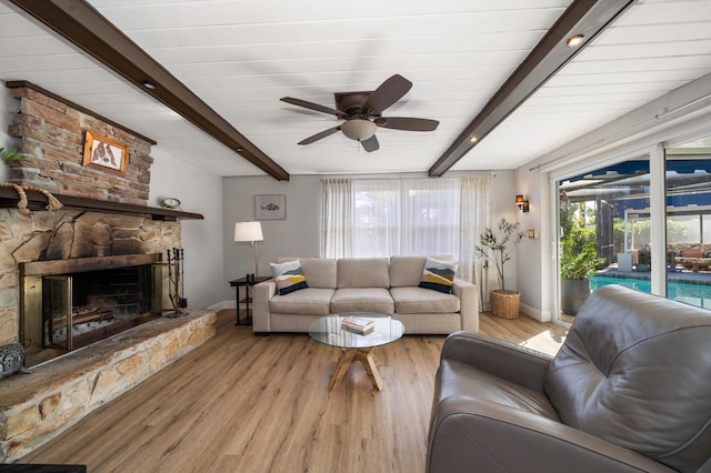 living room with beamed ceiling, ceiling fan, a stone fireplace, and light wood-type flooring