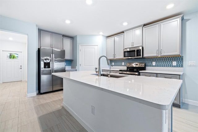 kitchen with sink, tasteful backsplash, appliances with stainless steel finishes, light stone countertops, and a kitchen island with sink