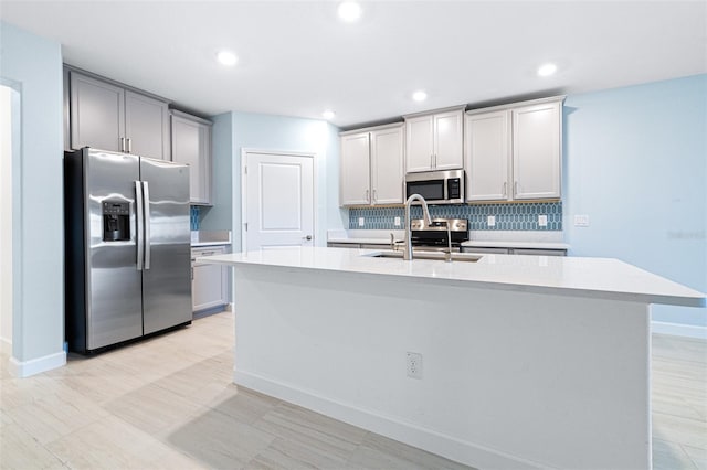 kitchen with sink, appliances with stainless steel finishes, gray cabinets, an island with sink, and backsplash