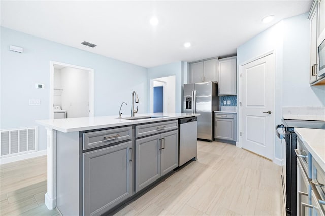 kitchen featuring sink, gray cabinetry, light hardwood / wood-style floors, stainless steel appliances, and a center island with sink