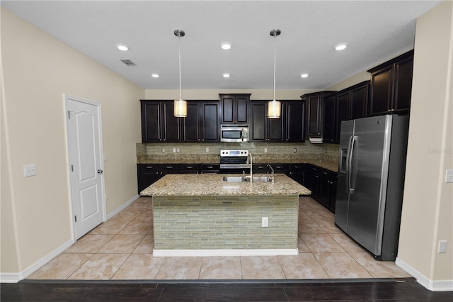 kitchen featuring sink, hanging light fixtures, appliances with stainless steel finishes, light stone countertops, and a kitchen island with sink