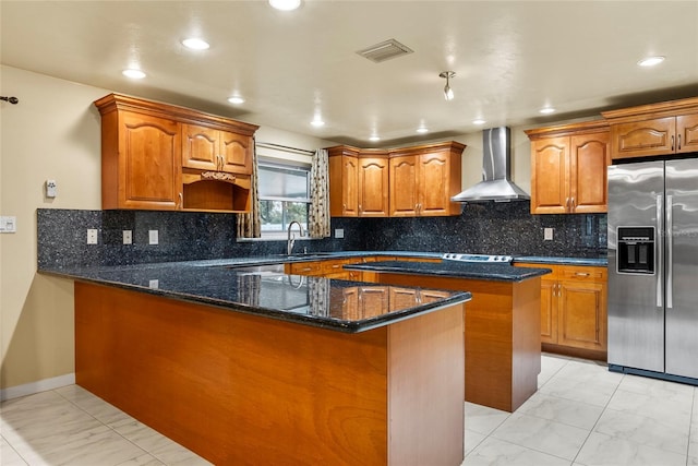 kitchen featuring sink, stainless steel fridge with ice dispenser, a kitchen island, dark stone counters, and wall chimney range hood