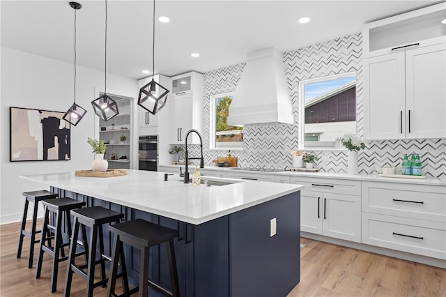 kitchen featuring sink, hanging light fixtures, white cabinets, a center island with sink, and custom exhaust hood