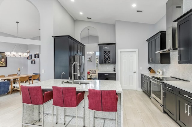 kitchen featuring appliances with stainless steel finishes, a breakfast bar area, hanging light fixtures, a center island with sink, and wall chimney exhaust hood