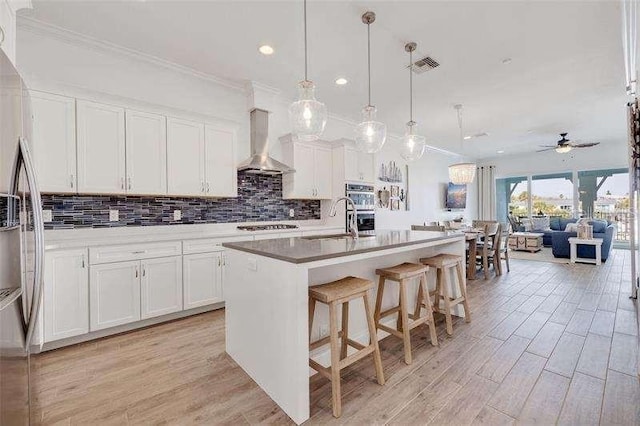 kitchen with pendant lighting, white cabinets, a kitchen island with sink, and wall chimney range hood