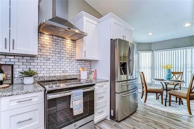 kitchen with white cabinets, decorative backsplash, light stone counters, stainless steel appliances, and wall chimney range hood