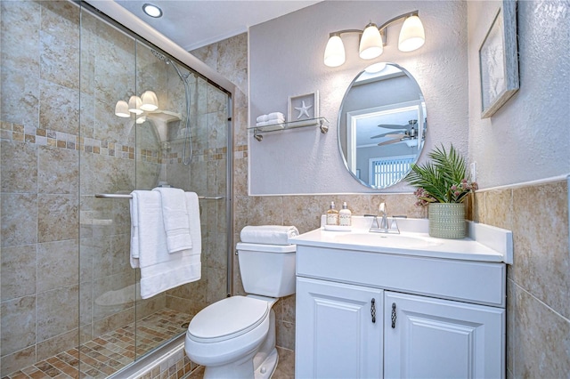 bathroom featuring a shower with door, ceiling fan, tile walls, vanity, and toilet