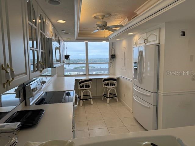 kitchen featuring light tile patterned floors, ceiling fan, white refrigerator with ice dispenser, white cabinets, and a raised ceiling