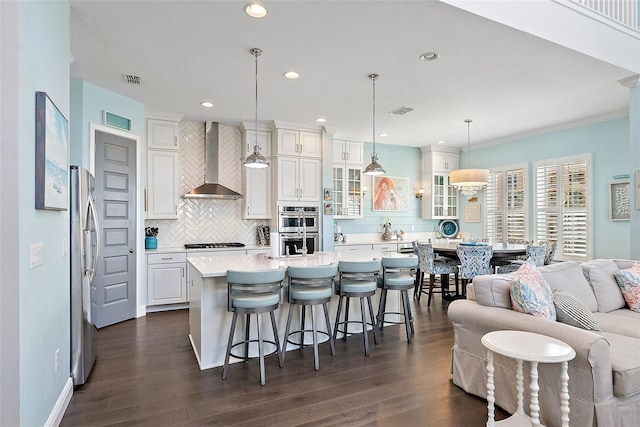 kitchen featuring appliances with stainless steel finishes, an island with sink, pendant lighting, wall chimney range hood, and white cabinets