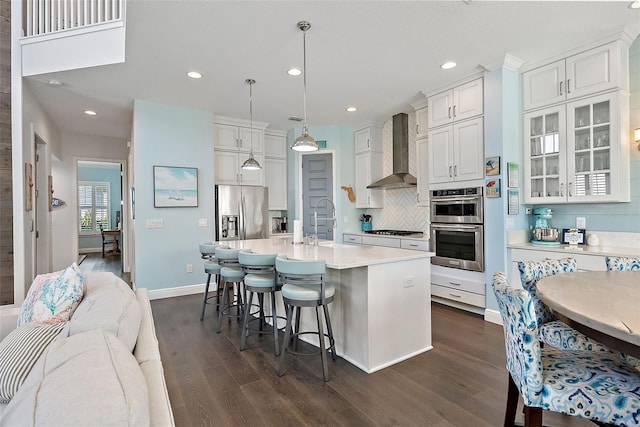 kitchen with white cabinetry, pendant lighting, stainless steel appliances, a kitchen island with sink, and wall chimney range hood