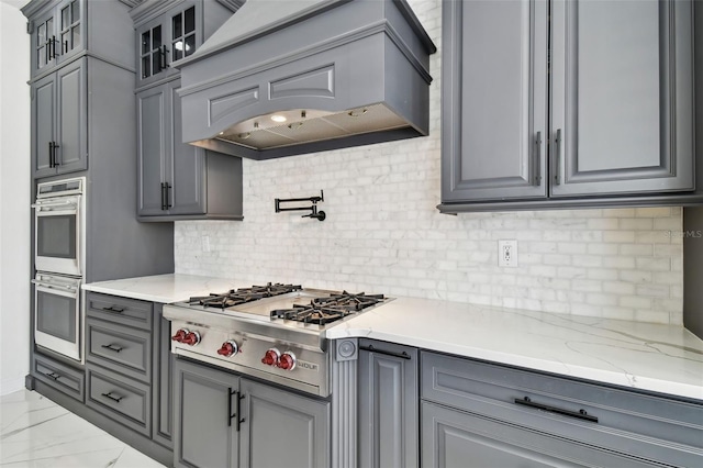 kitchen with premium range hood, gray cabinetry, stainless steel appliances, light stone countertops, and backsplash