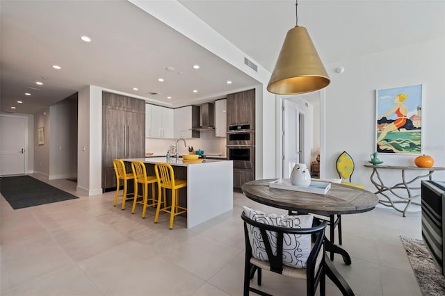 kitchen with wall chimney range hood, a breakfast bar, double oven, hanging light fixtures, and an island with sink