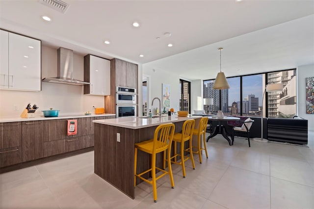 kitchen featuring double oven, an island with sink, white cabinets, decorative light fixtures, and wall chimney exhaust hood