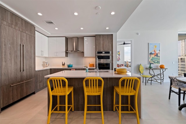 kitchen featuring wall chimney range hood, a breakfast bar area, white cabinetry, a center island with sink, and stainless steel double oven