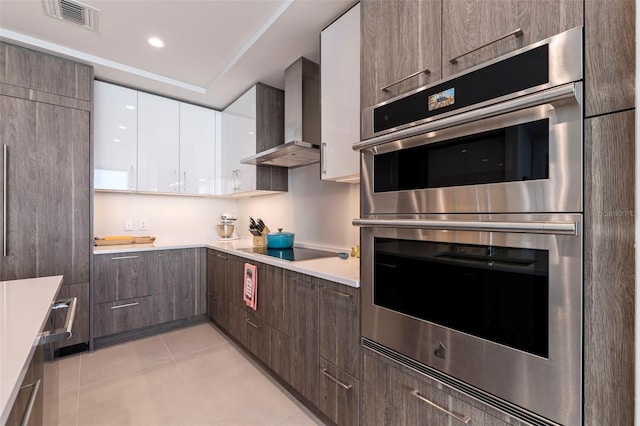 kitchen featuring wall chimney exhaust hood, light tile patterned floors, black electric cooktop, stainless steel double oven, and white cabinets