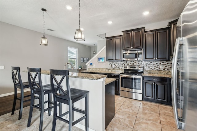 kitchen featuring hanging light fixtures, a kitchen breakfast bar, stainless steel appliances, light stone countertops, and a kitchen island with sink