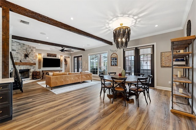 dining space featuring hardwood / wood-style flooring, ceiling fan with notable chandelier, beam ceiling, ornamental molding, and a stone fireplace