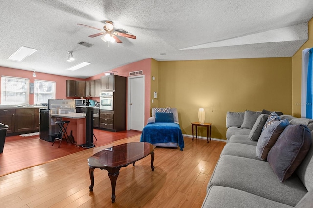 living room featuring ceiling fan, a textured ceiling, vaulted ceiling with skylight, and light wood-type flooring