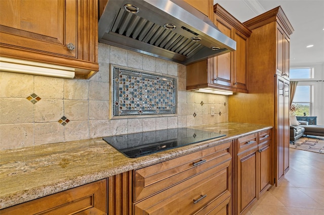 kitchen with wall chimney range hood, black electric stovetop, light stone countertops, light tile patterned flooring, and decorative backsplash