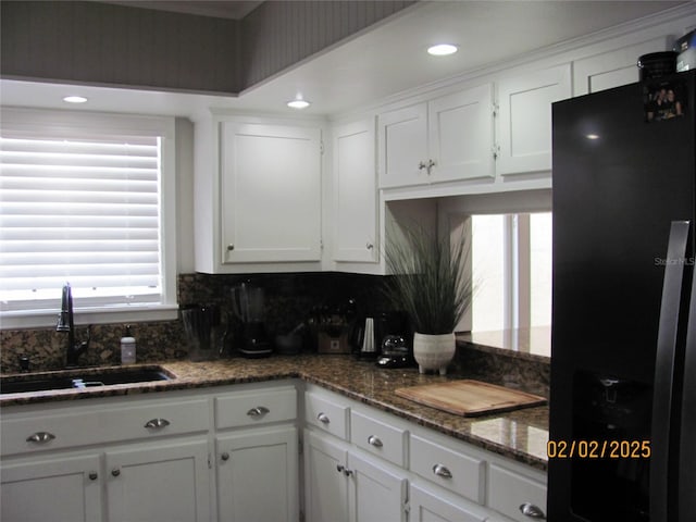 kitchen featuring sink, dark stone countertops, black refrigerator, white cabinets, and backsplash