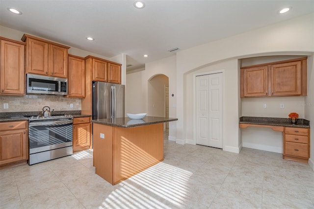 kitchen featuring appliances with stainless steel finishes, built in desk, decorative backsplash, dark stone counters, and a center island