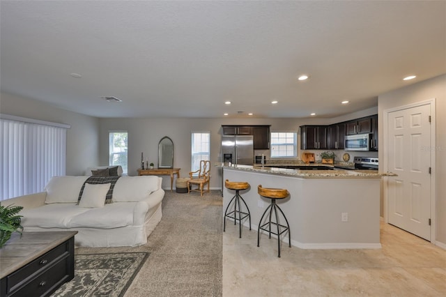 kitchen featuring appliances with stainless steel finishes, a breakfast bar, a kitchen island with sink, dark brown cabinetry, and a healthy amount of sunlight